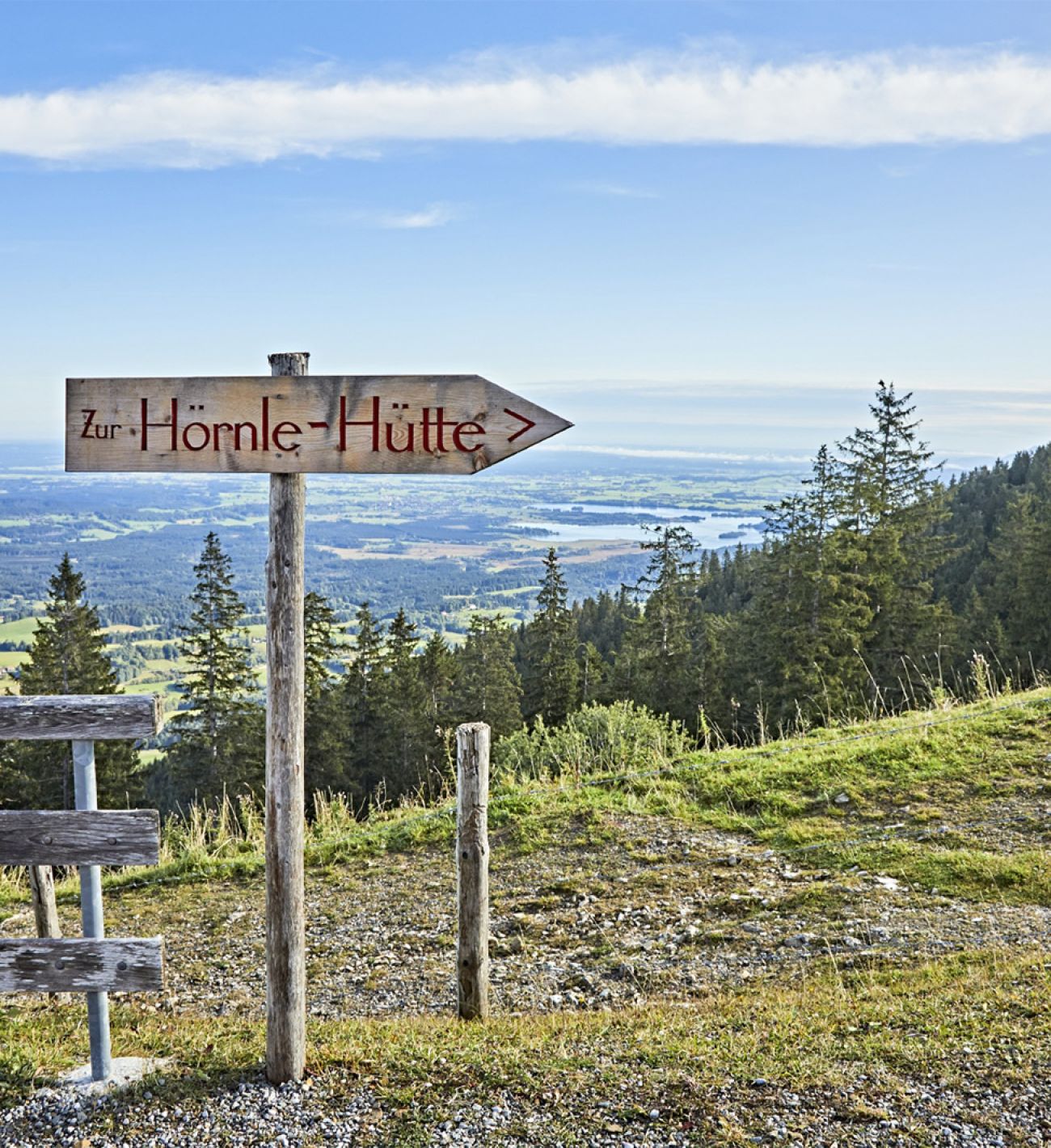 Anreise Das Bild zeigt einen hölzernen Wegweiser mit der Aufschrift „Zur Hörnle-Hütte“ an einem Berghang mit Blick auf eine weitläufige, hügelige Landschaft mit Wäldern und Seen. Im Vordergrund befindet sich ein einfacher Holzzaun. Der Himmel ist klar und blau mit ein paar wenigen Wolken. Das Bild strahlt Ruhe und die Schönheit der Natur aus, und lädt zum Wandern ein.