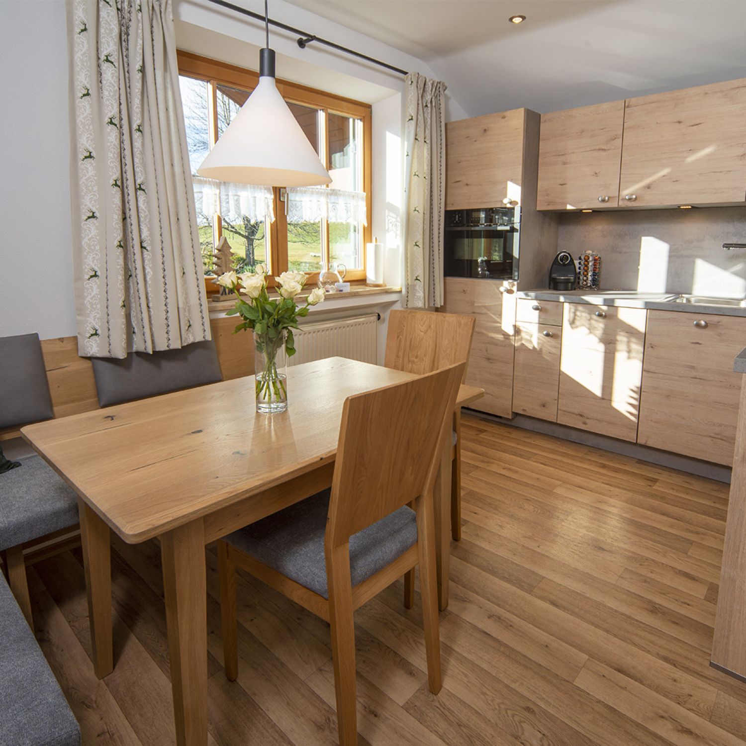 Das Bild zeigt eine helle, moderne Küche mit integrierter Essecke. Die Küche ist in hellen Holztönen gehalten und verfügt über moderne Geräte. Ein Esstisch aus hellem Holz steht im Vordergrund, umgeben von Stühlen mit grauen Stoffbezügen. Eine Eckbank mit grauen Kissen ist ebenfalls vorhanden. Ein Fenster mit hellen Vorhängen lässt viel Tageslicht in den Raum. Auf dem Tisch steht eine Vase mit weißen Rosen. Die Atmosphäre ist freundlich und einladend.